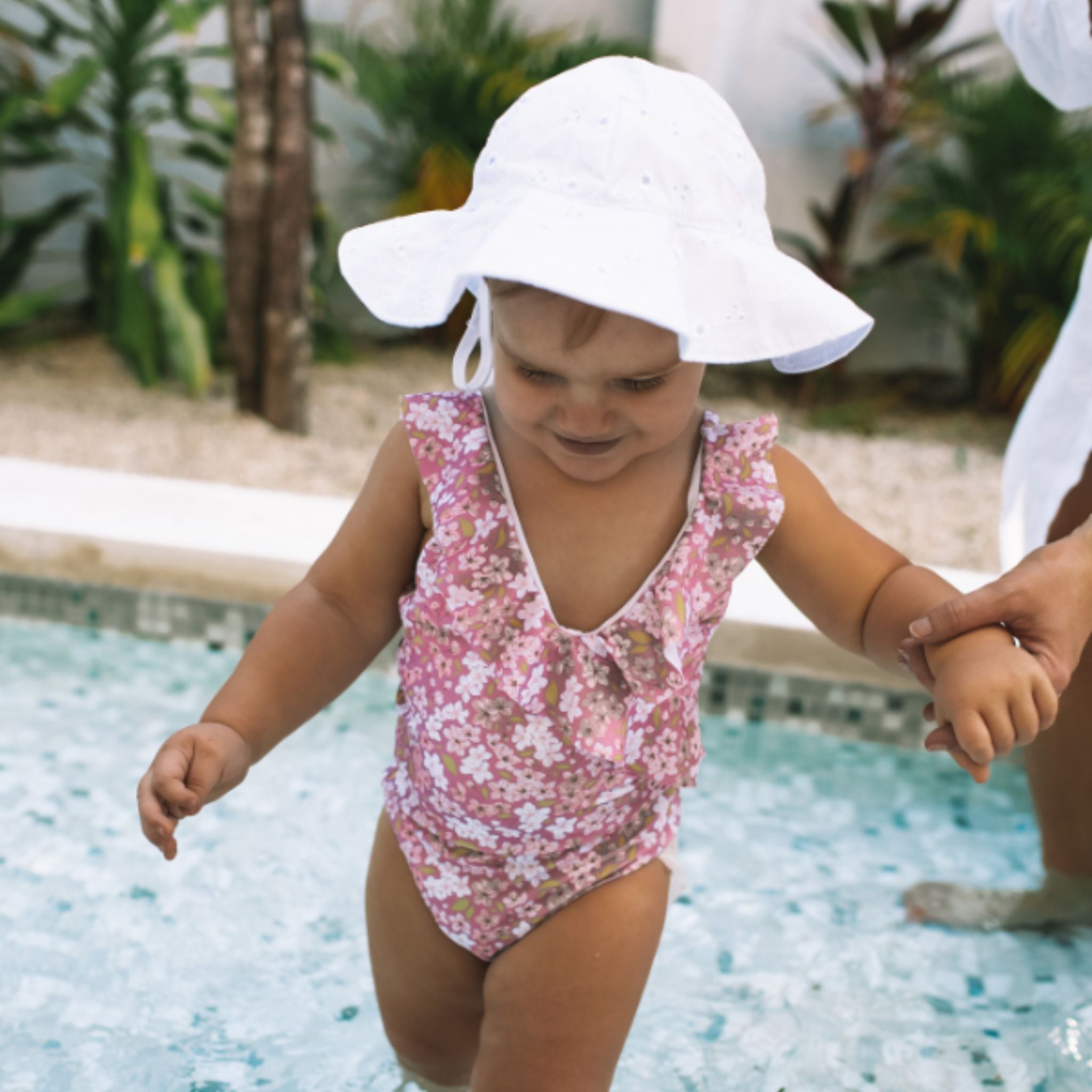 Child in a floral swimsuit and sun hat walking by a pool with a person holding their hand.