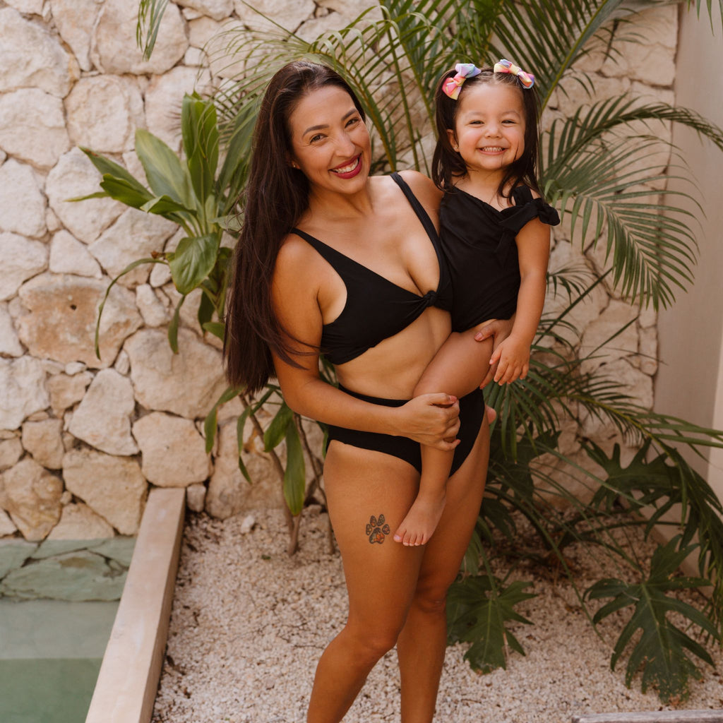 Woman and child in black swimwear standing in front of a stone wall with plants.