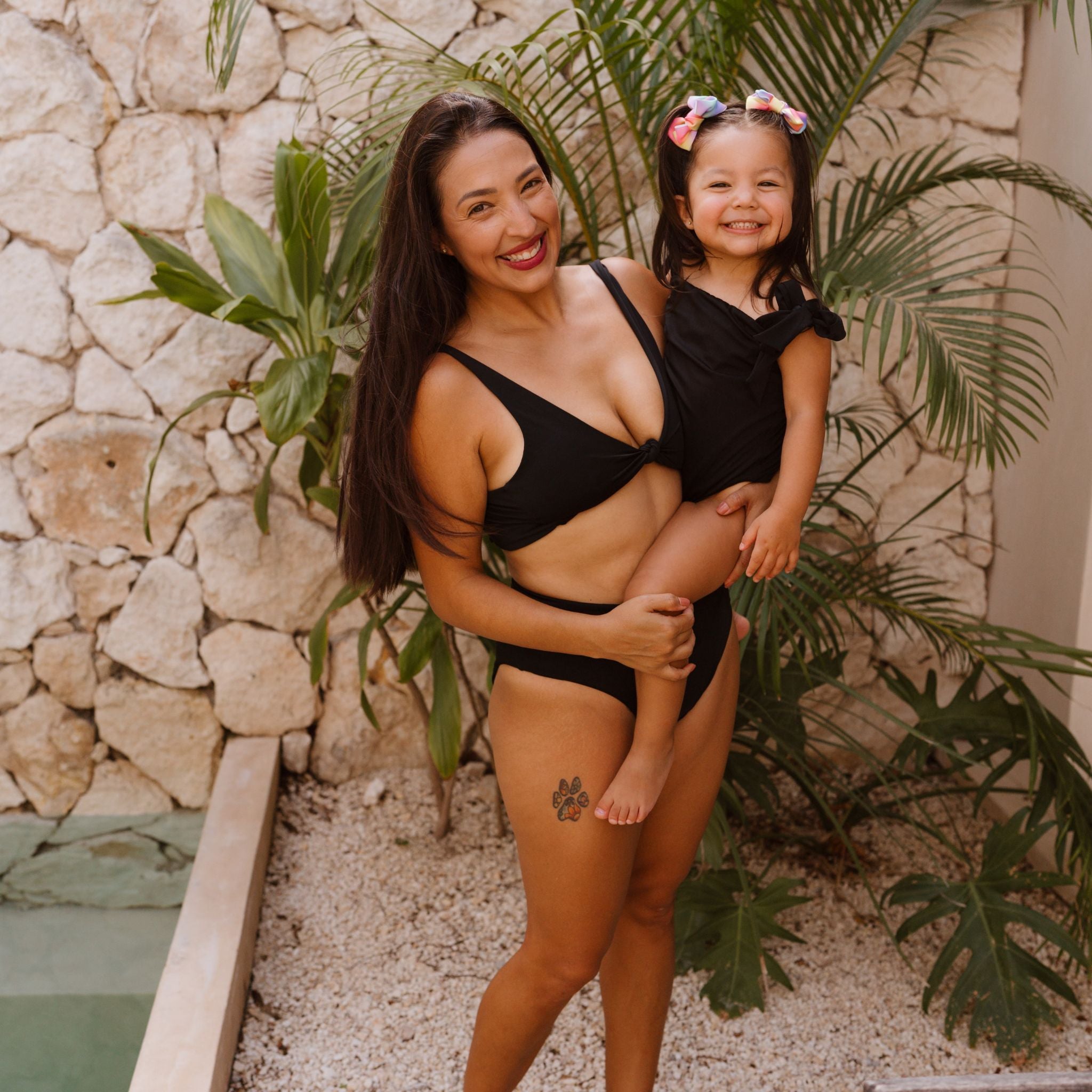 Woman and child in black swimwear standing in front of a stone wall with plants.