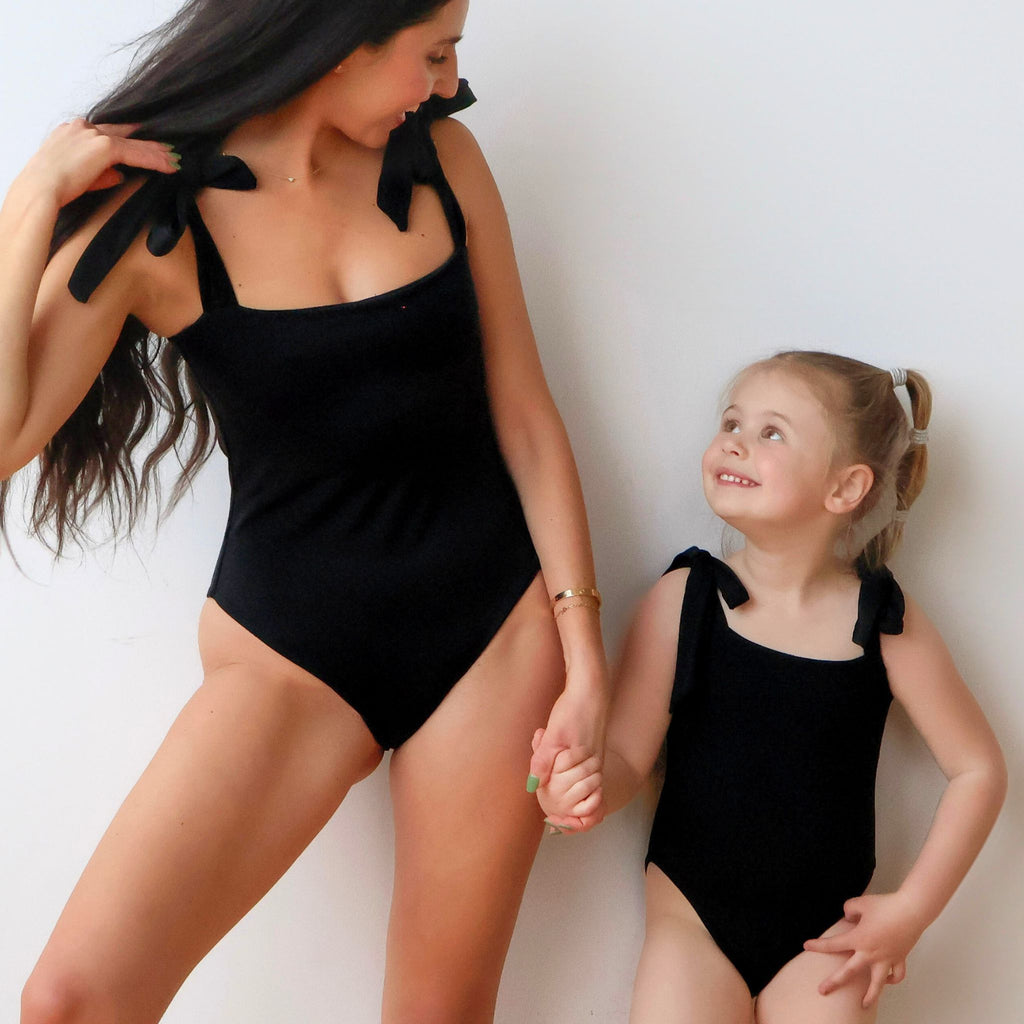 Woman and young girl in matching black swimsuits against a white background