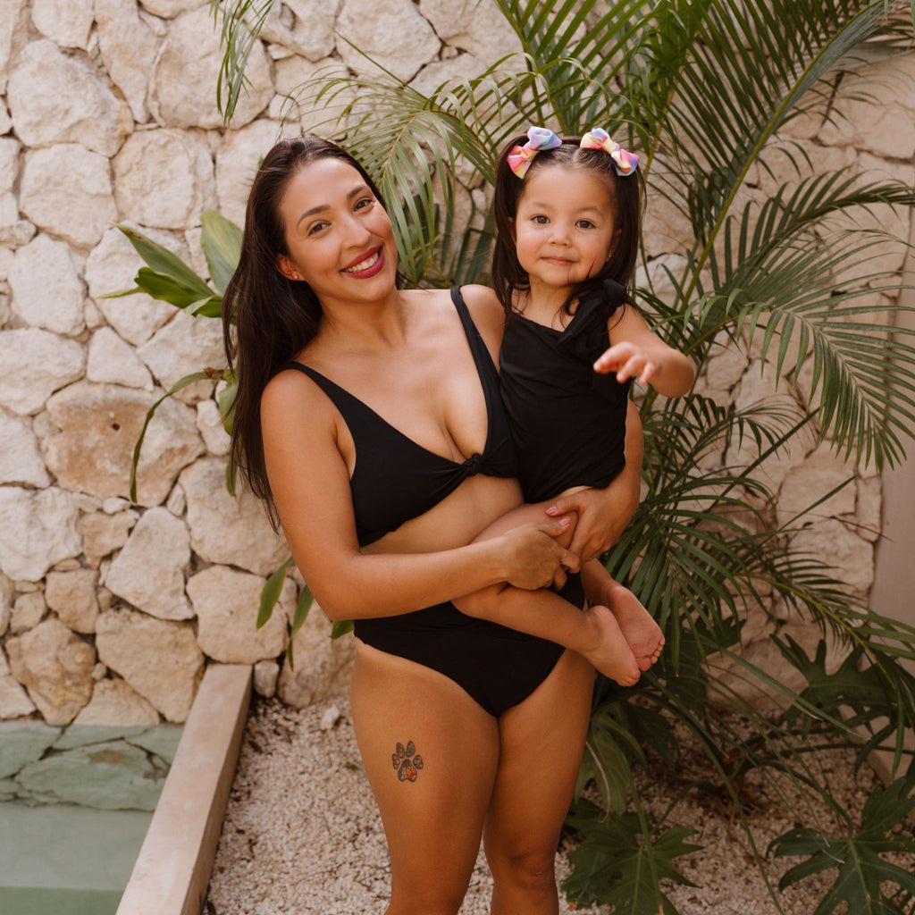 Woman and child in matching black swimsuits standing against a stone wall with greenery.